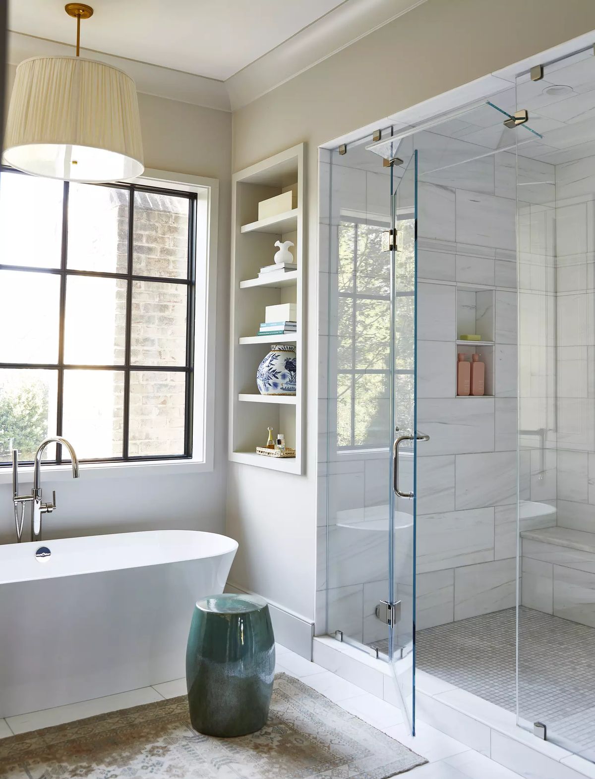 Neutral luxury bathroom with freestanding soaking tub, glass marble shower, built-in shelves, and black framed window.