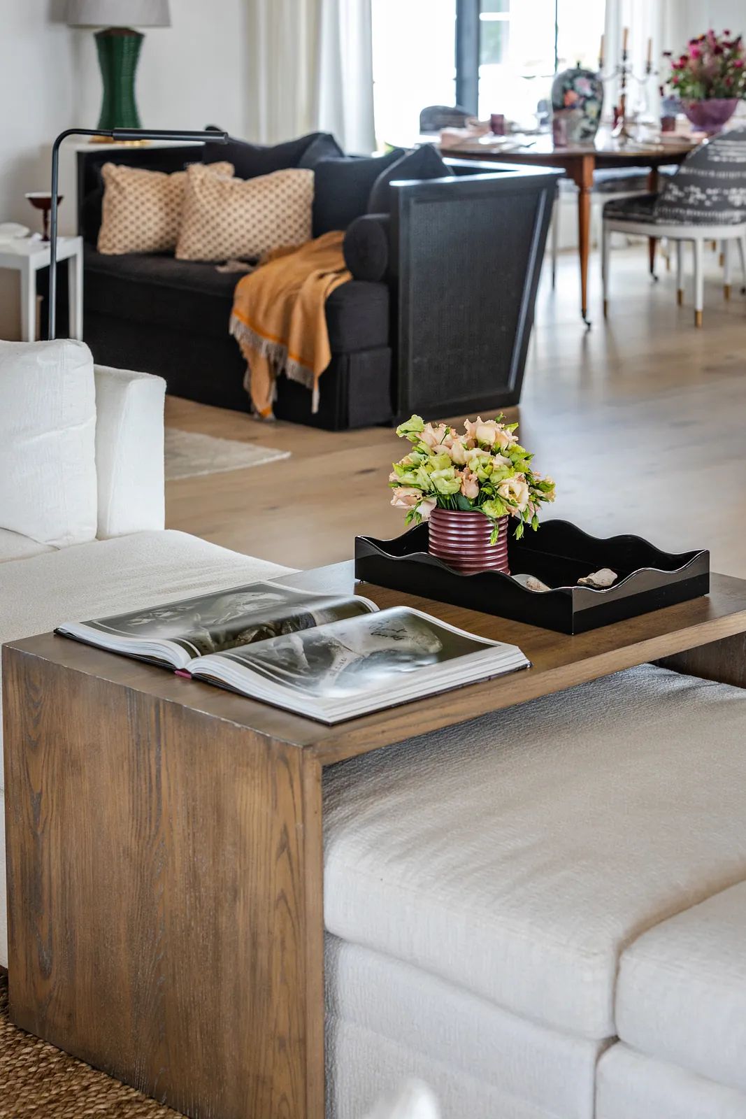 Living room with white sectional, dark accent sofa, wooden side table, flowers, and large windows.