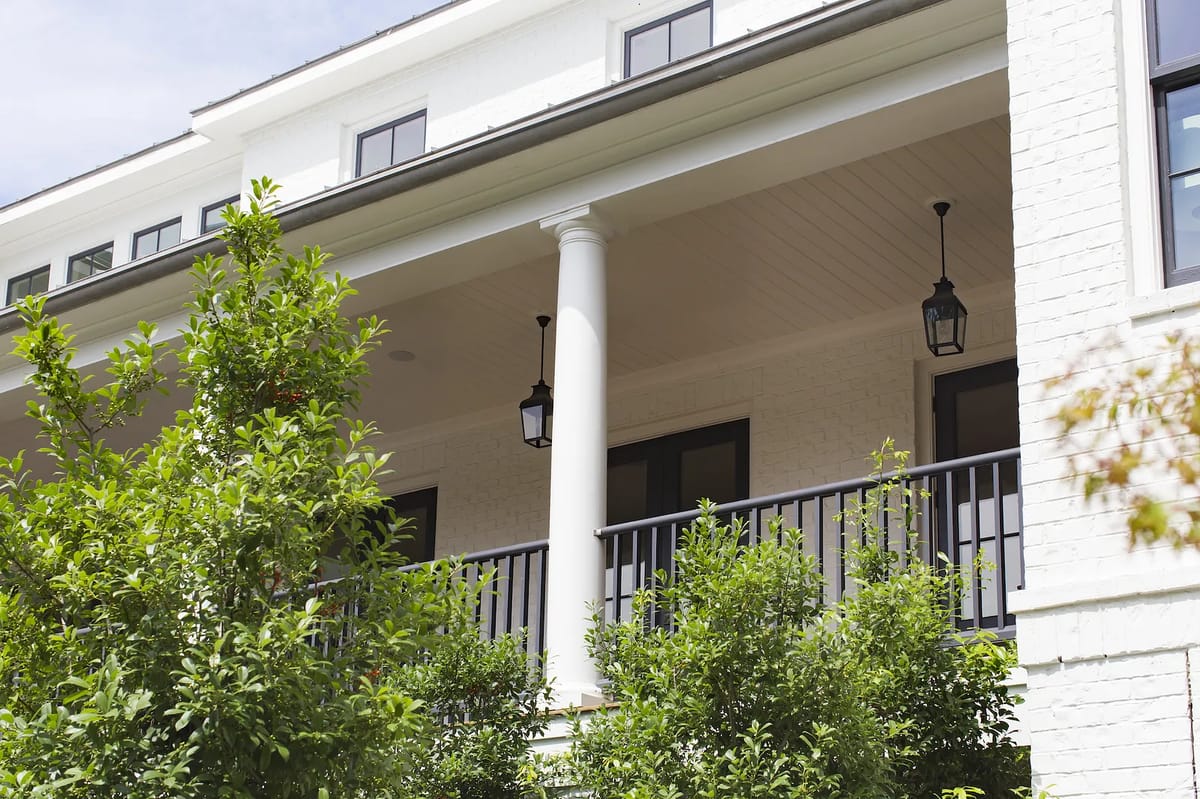 Exterior front view with covered porch, columns, railing, lanterns, and surrounding greenery.