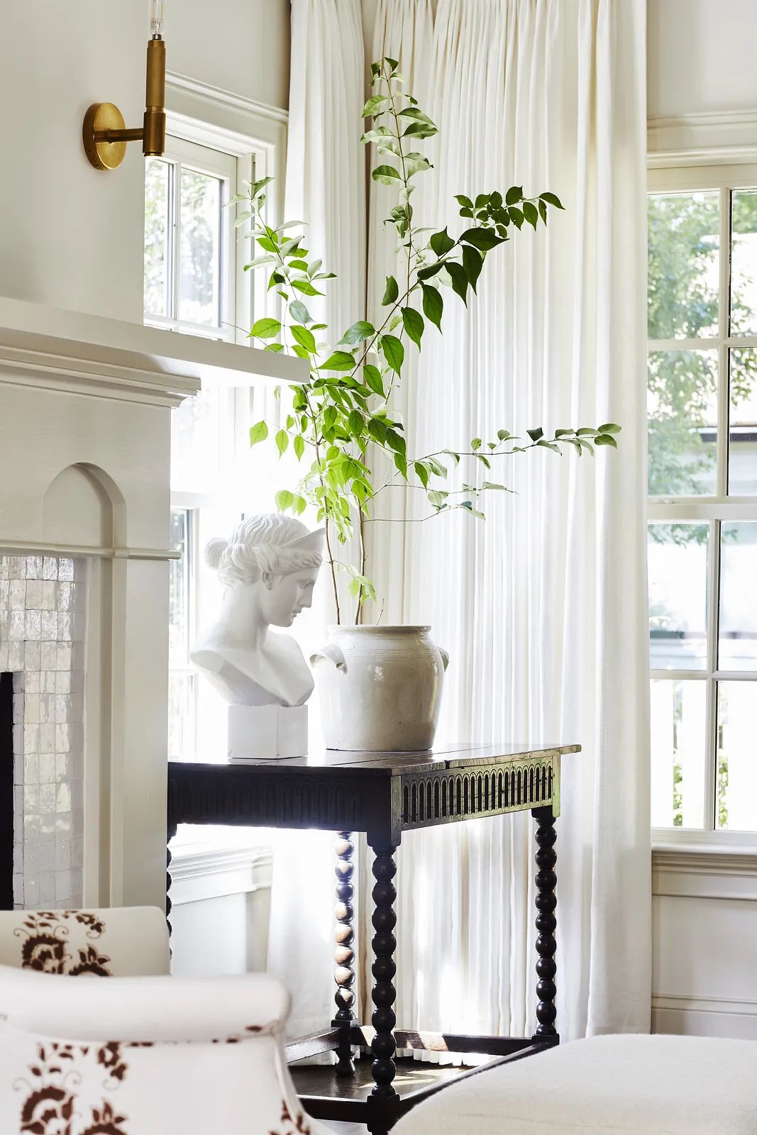 Living room with side table, bust sculpture, ceramic pot, and sheer curtains by large windows.