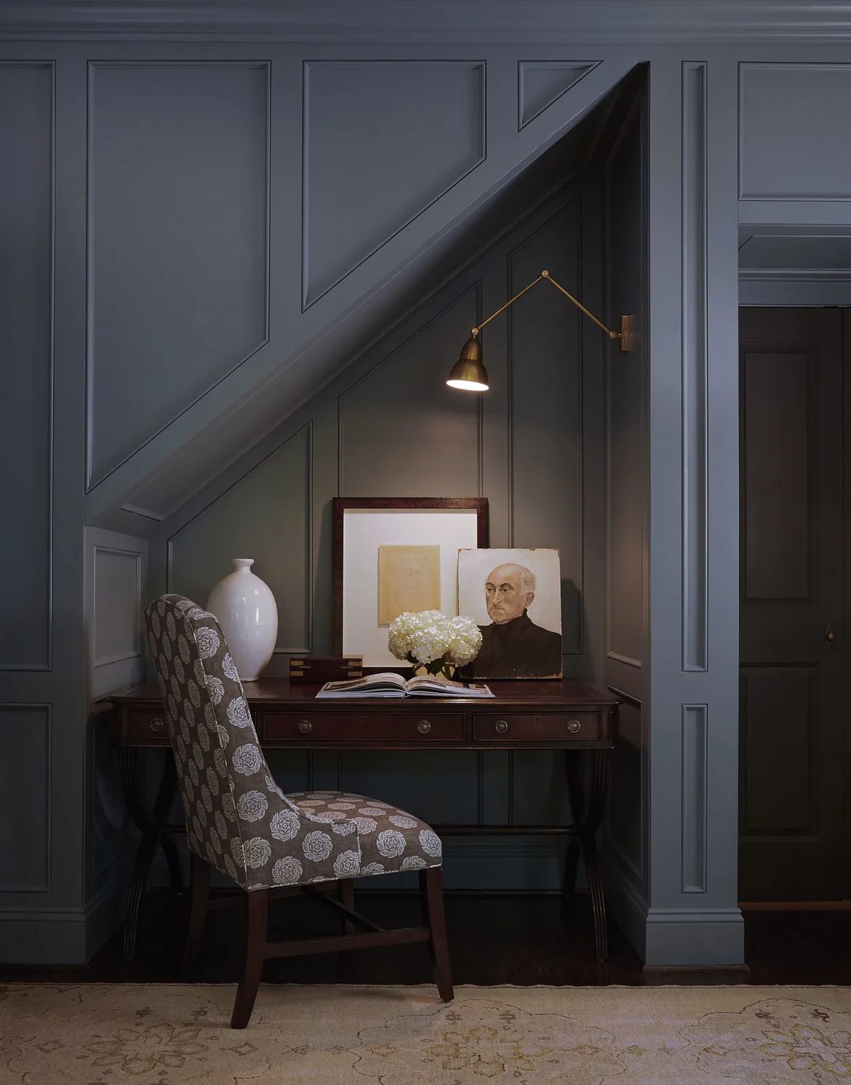 Office with dark blue panels, wooden desk, chair, flowers, artwork, and wall-mounted light fixture