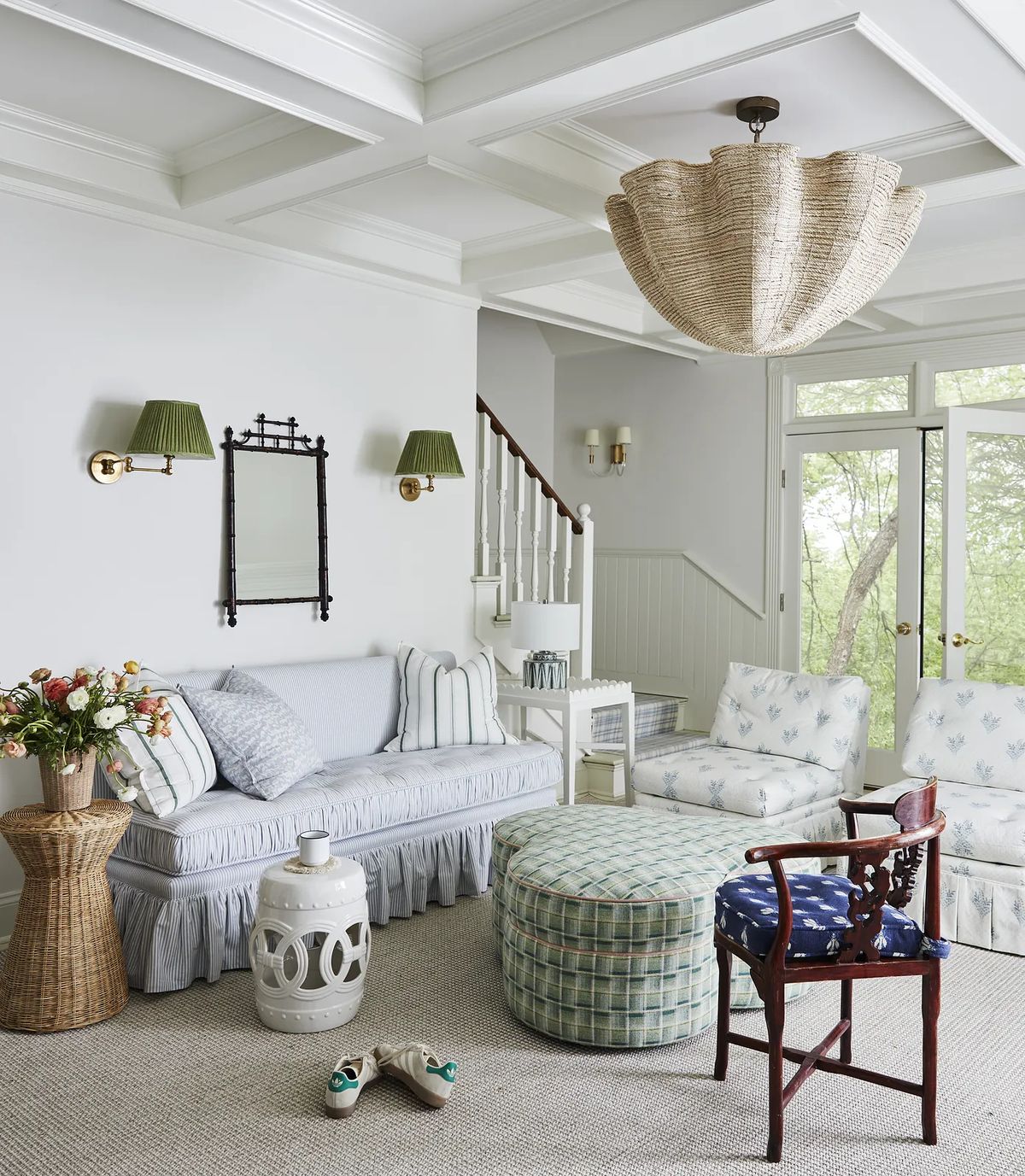 Living room with blue sofa, patterned ottoman, chairs, floral arrangement, coffered ceilings, and large windows.