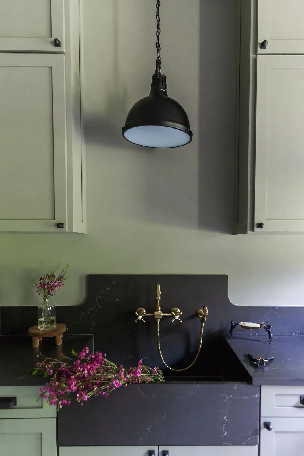 Kitchen with black countertop, grey cabinets, wall-mounted sink, brass faucet, and black pendant light