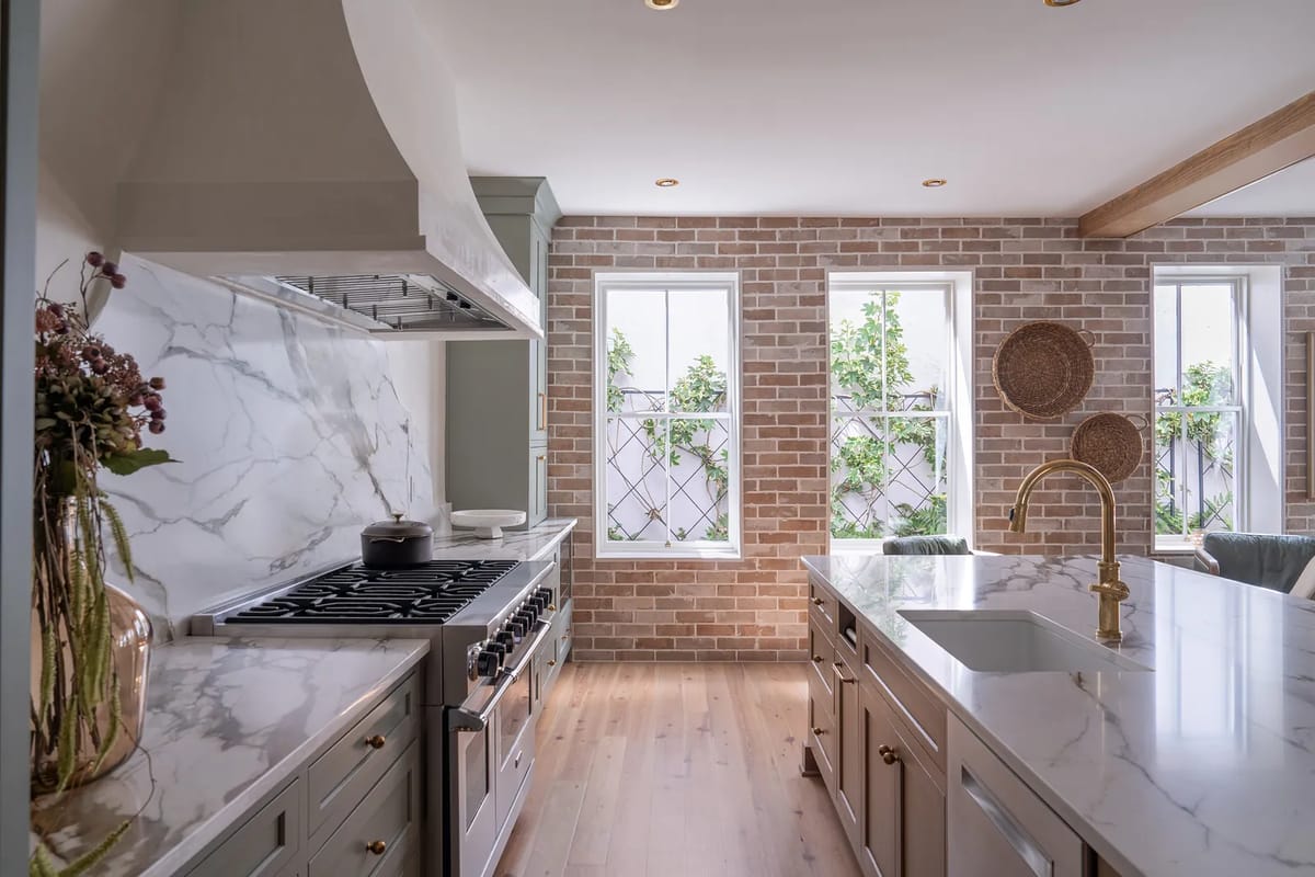Kitchen with marble-topped island, gas stove, exposed brick walls, and large windows with greenery view.