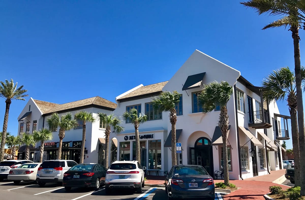 Exterior view of a retail plaza with white buildings, palm trees, and visible signage.
