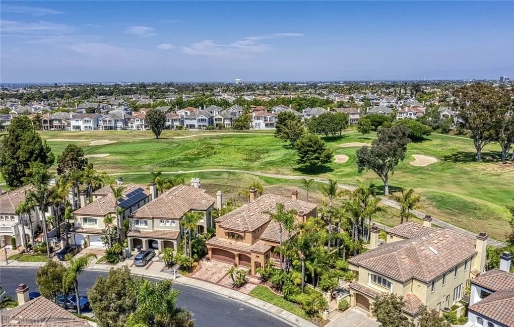 Aerial view of suburban homes with tiled roofs, lawns, palm trees, and a golf course in the background.