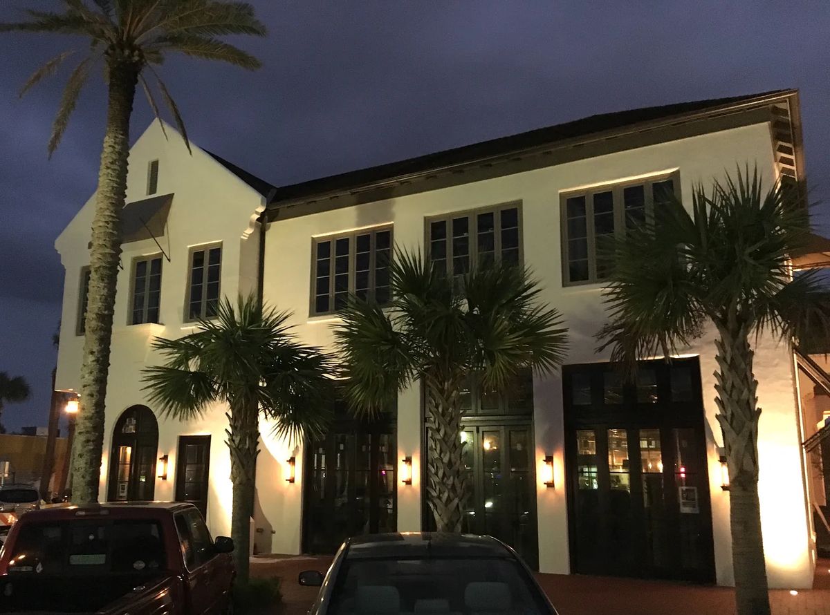 Exterior view of a building with large windows, palm trees, and wall-mounted lighting fixtures.