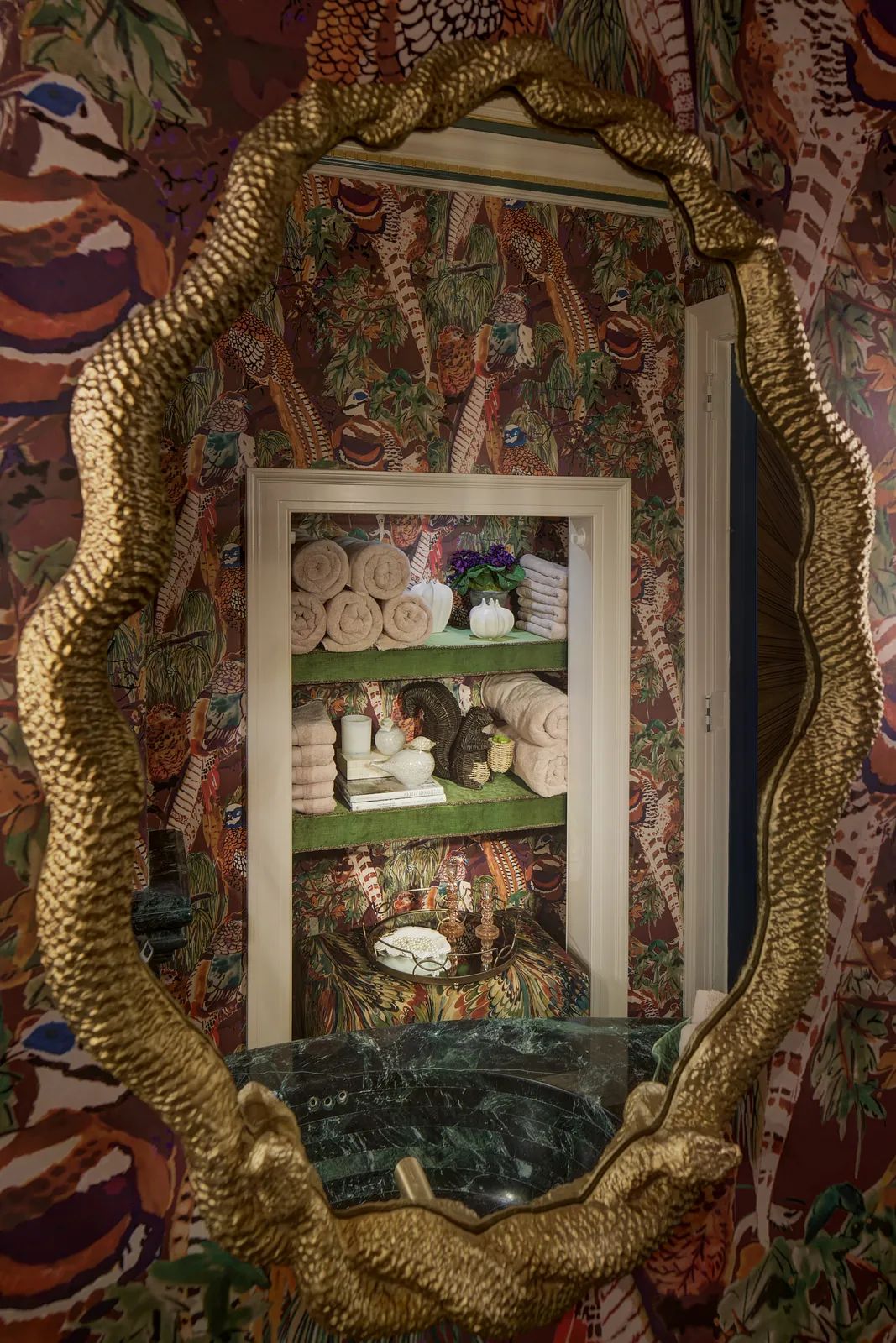 Powder room with gold-framed mirror, green marble countertop, floral wallpaper, and decorative towel shelf.