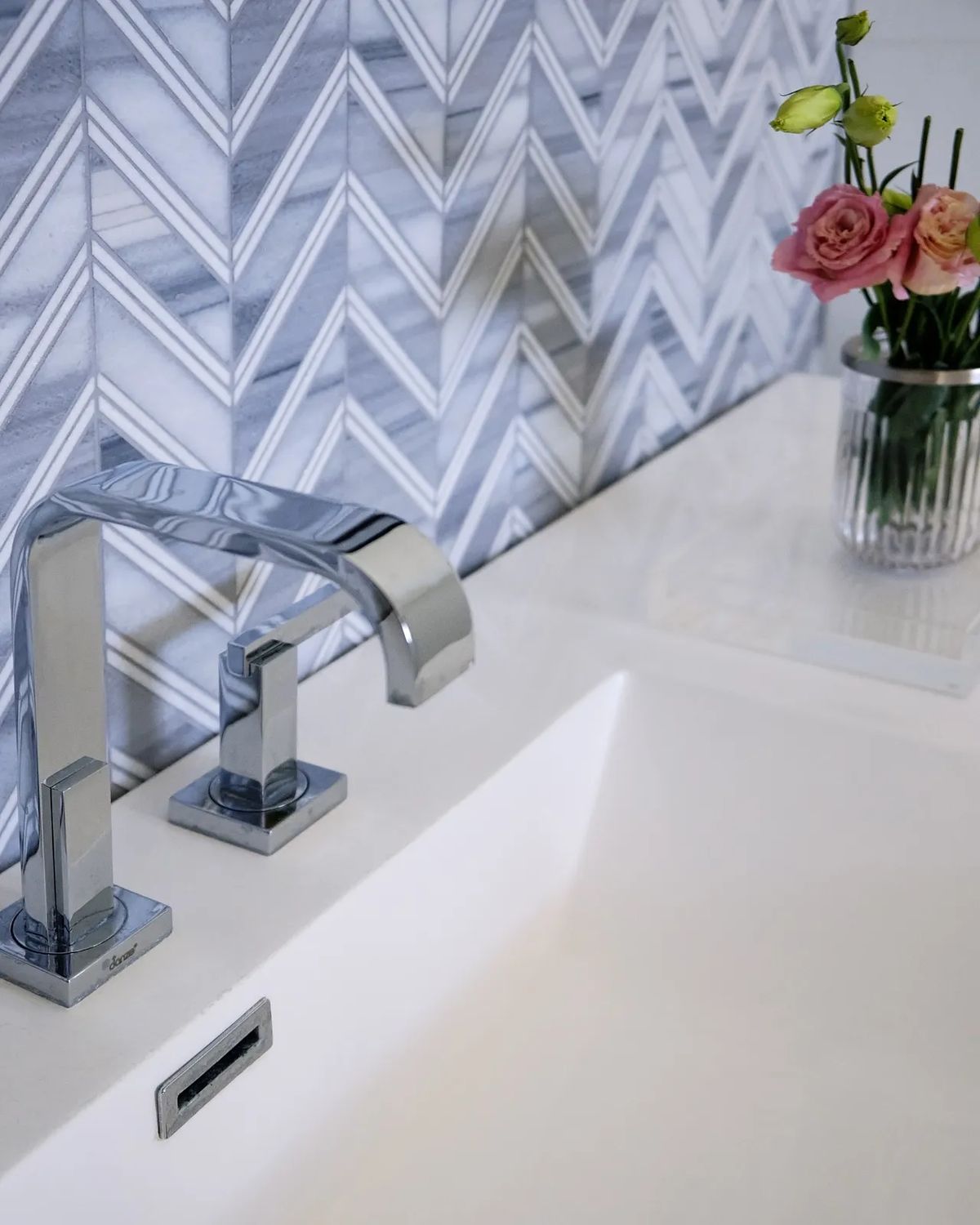 Bathroom with white sink, chrome faucet, chevron tile wall, and vase of pink roses