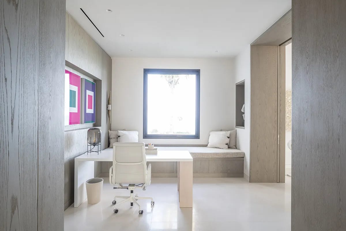 Office with white desk, ergonomic chair, built-in bench, cushions, artwork, and large window.
