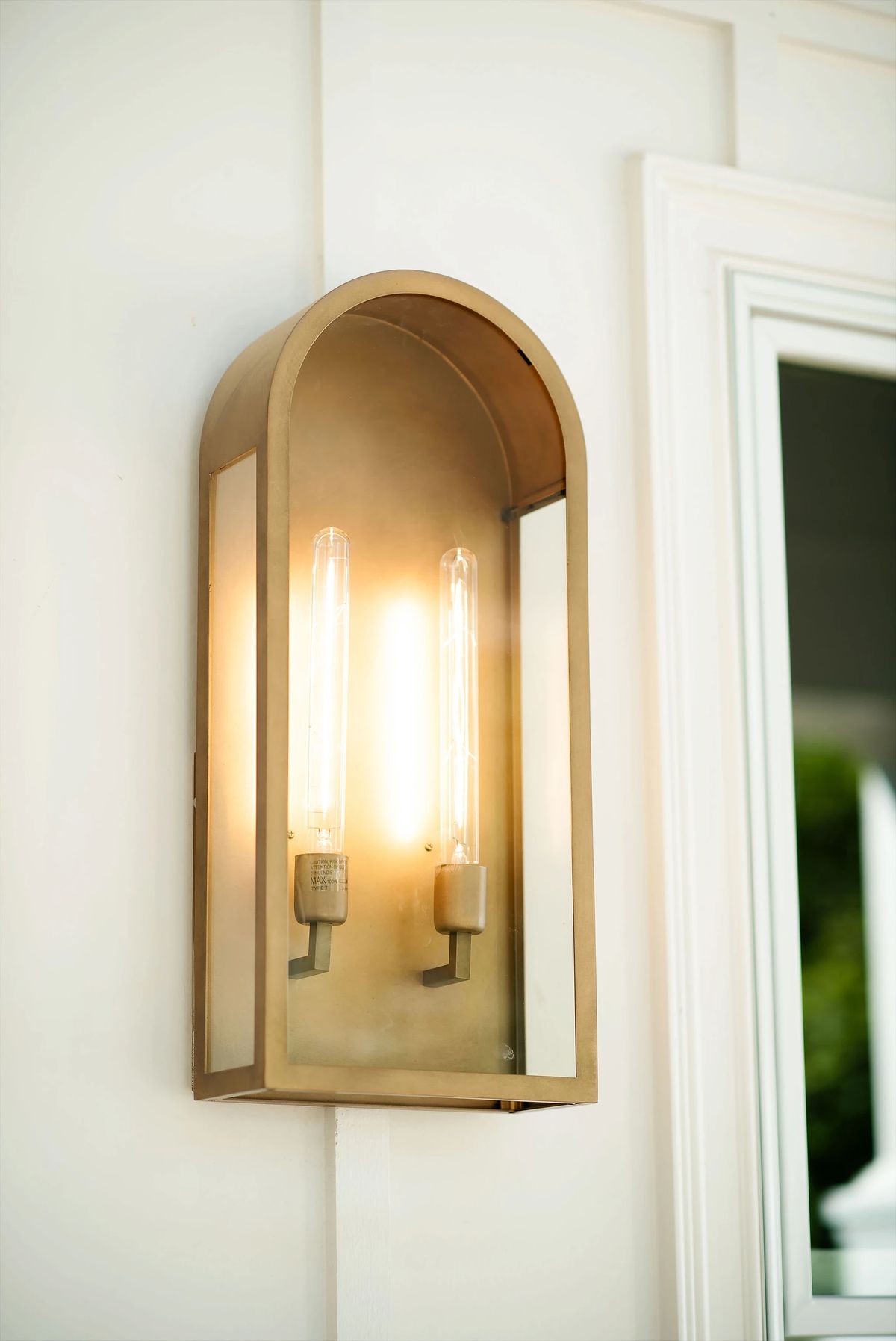 Wall-mounted light fixture with two exposed bulbs in gold frame near a large window.
