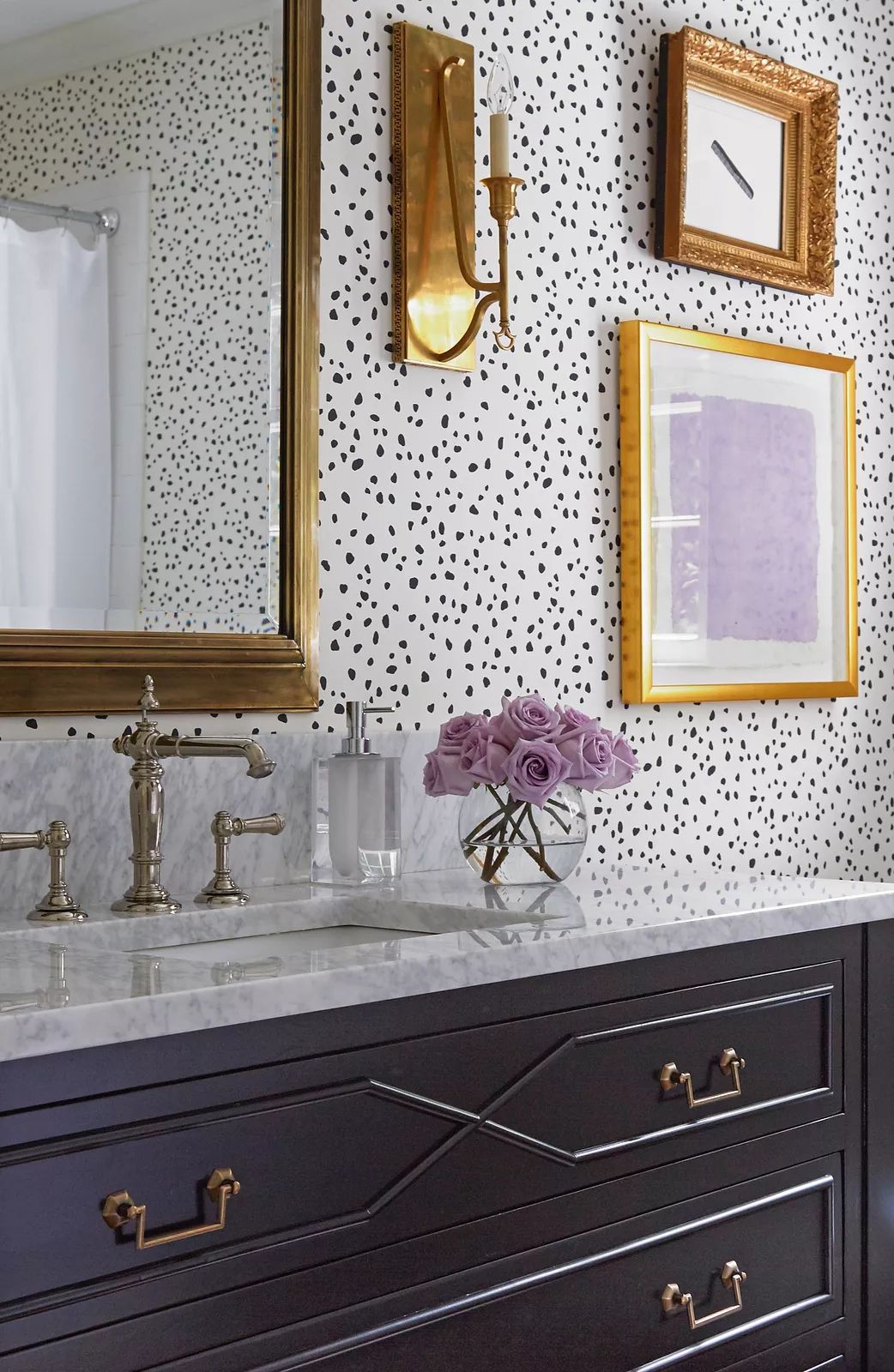 Bathroom with dark wood vanity, marble countertop, gold-framed mirror, sconces, and polka dot wallpaper.