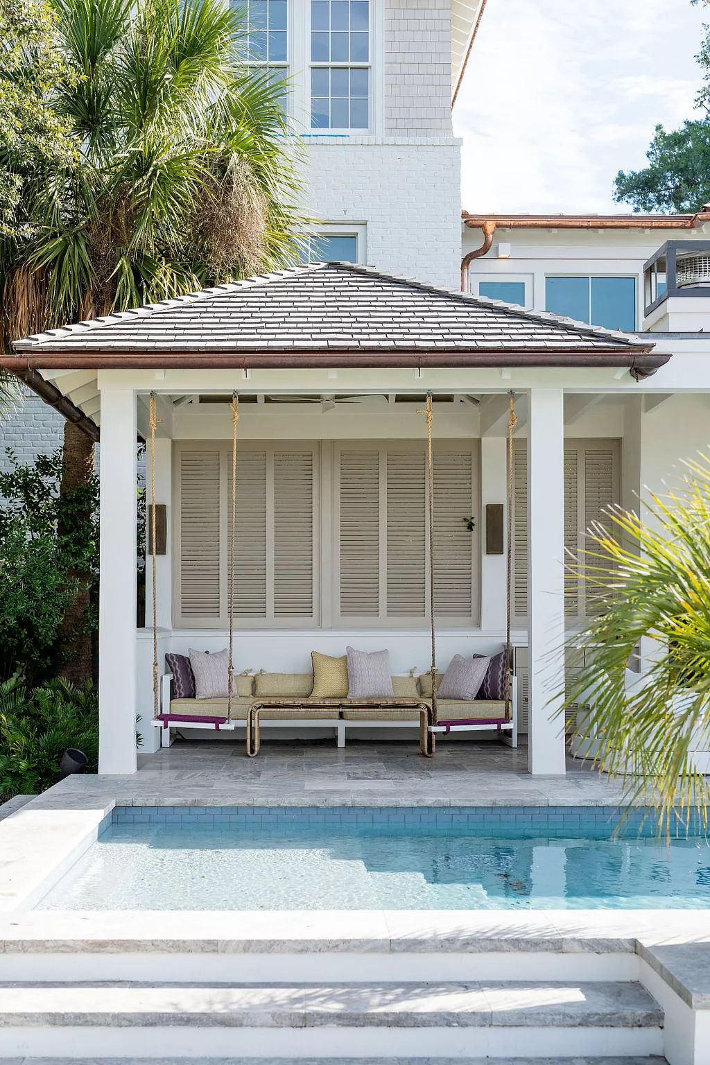 Patio with swing bench, colored pillows, surrounded by palm trees and a swimming pool under a gazebo.