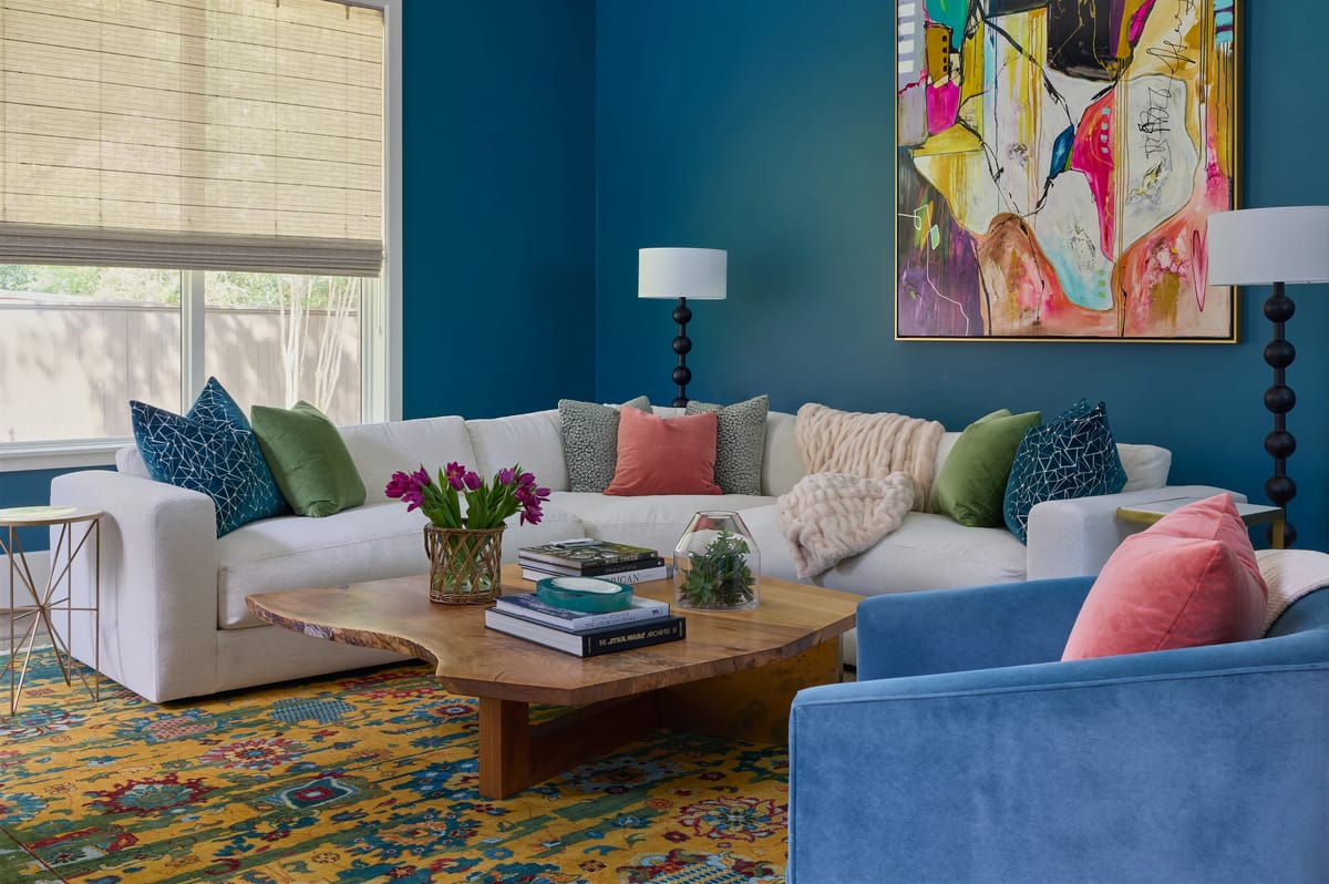 Living room with white sofa, blue armchair, wooden coffee table, colorful rug, and large windows.