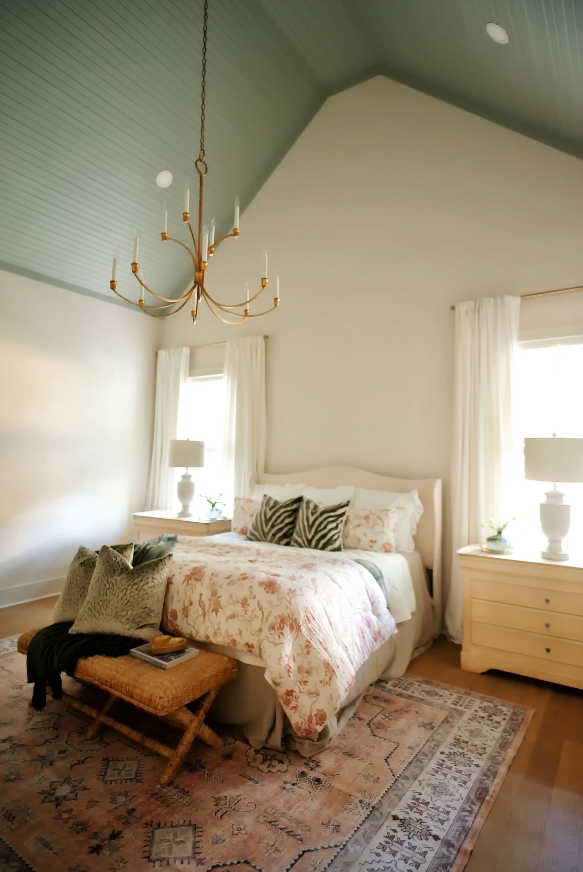 Bedroom with floral quilt, nightstands, lamps, woven bench, and large windows with sheer curtains