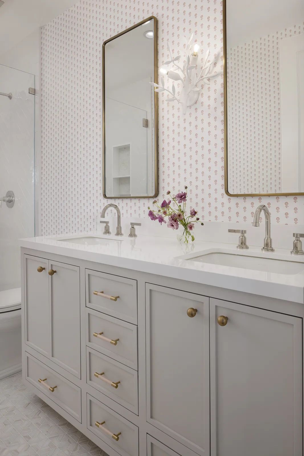 Bathroom with gray double vanity, white countertop, patterned wallpaper, and glass shower enclosure