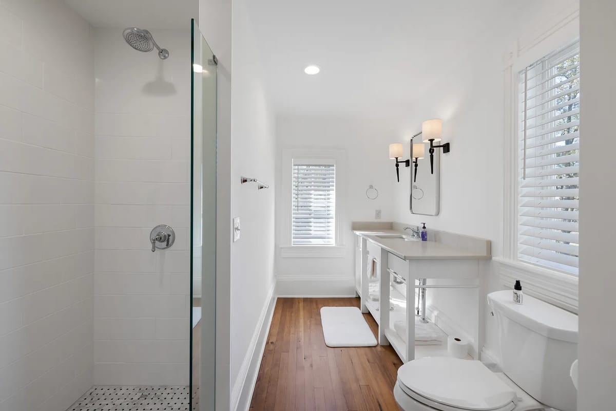 Bathroom with glass shower, white vanity, mirrors, wood floor, and window with blinds.