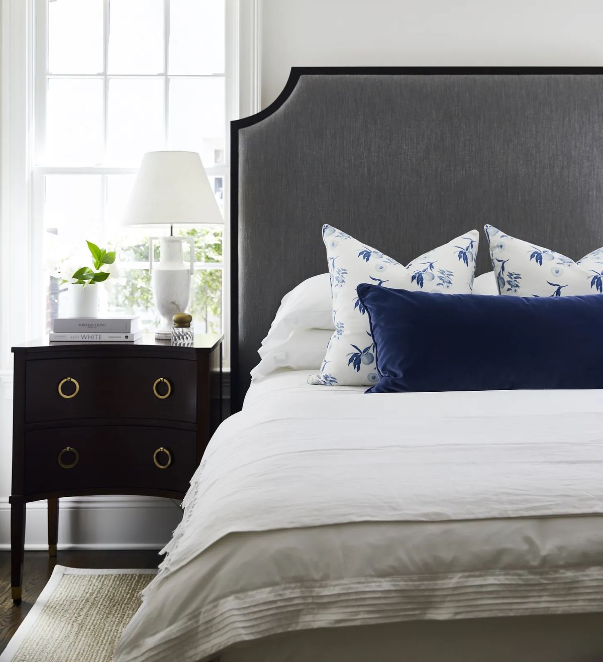 Bedroom with gray upholstered headboard, white bedding, nightstand, lamp, and window for natural light.