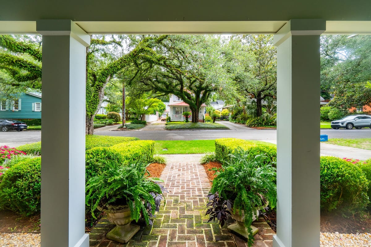 Exterior front view with brick pathway, green hedges, ferns, and white columns.