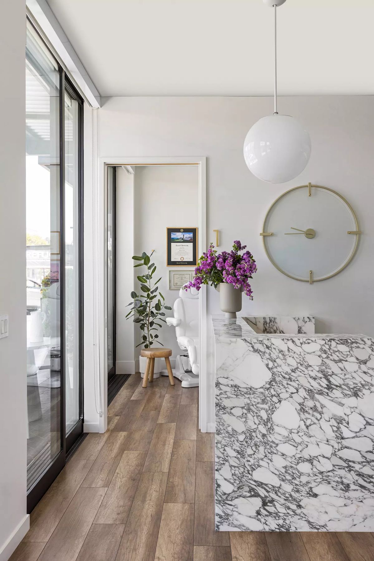 Foyer with marble reception desk, floral arrangement, wooden stool, wall clock, and tall glass doors.