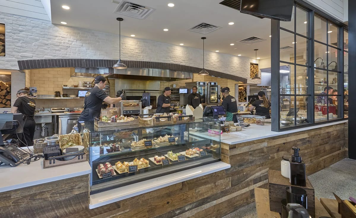 Kitchen with large counter, display case for pastries, employees, and brick and wood accents