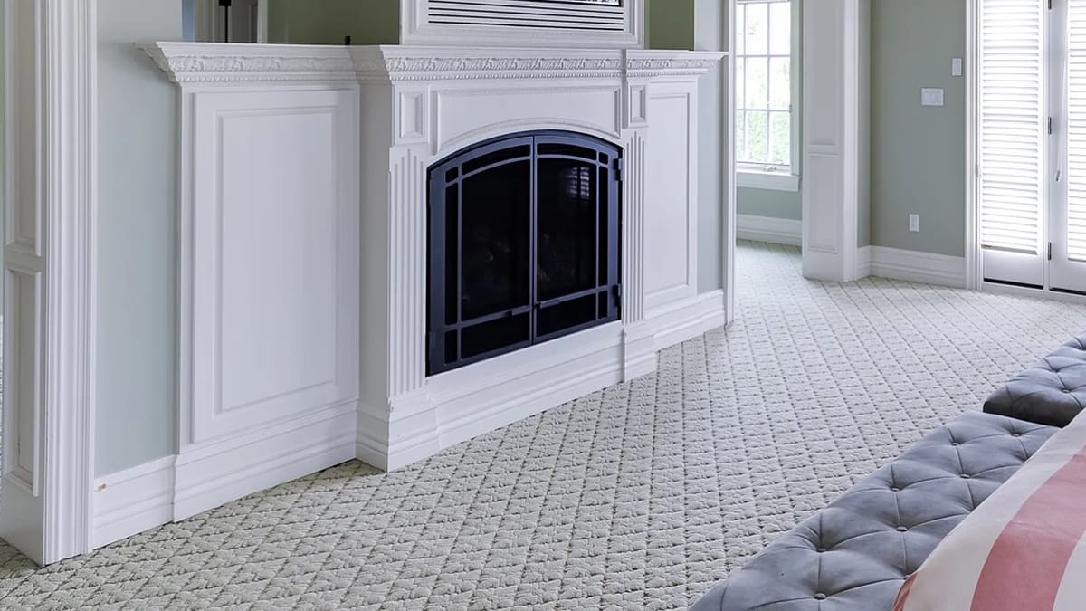 Living room with large fireplace, pale green walls, quilted rug, and windows with white shutters.