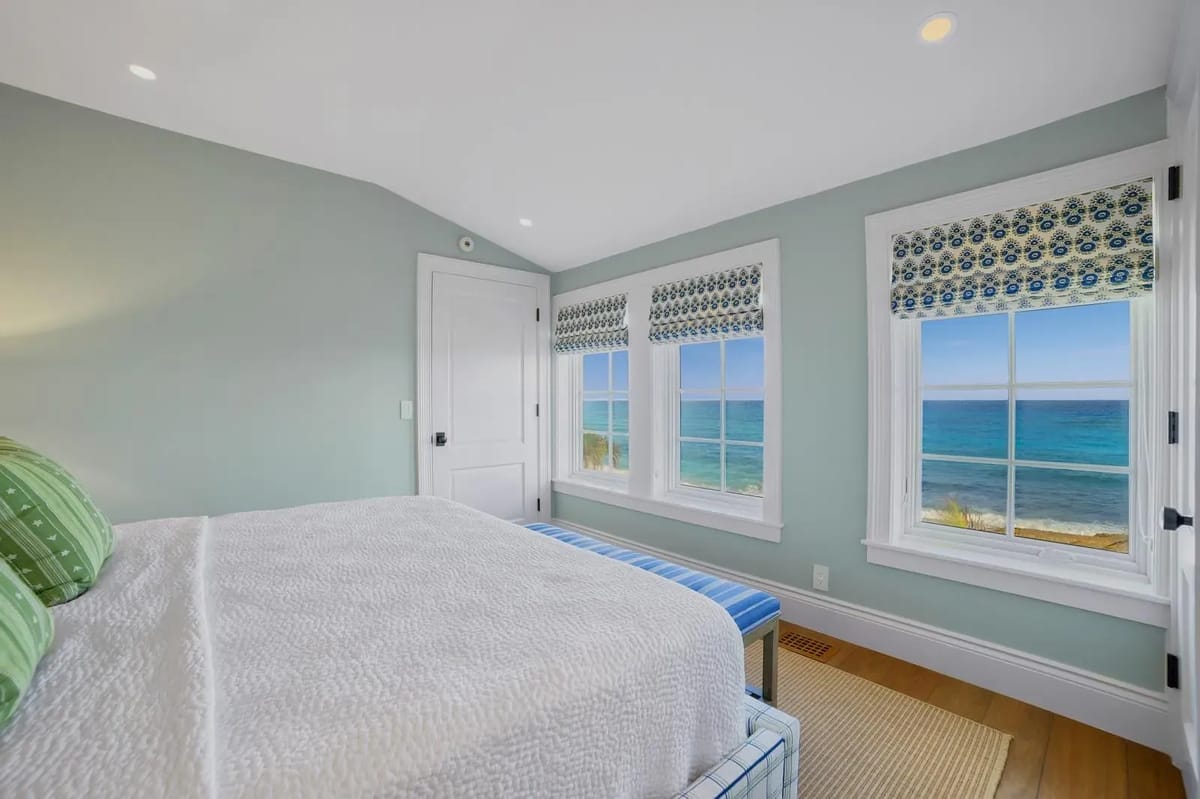 Bedroom with white quilted bed, striped cushions, blue bench, large windows, and light blue walls