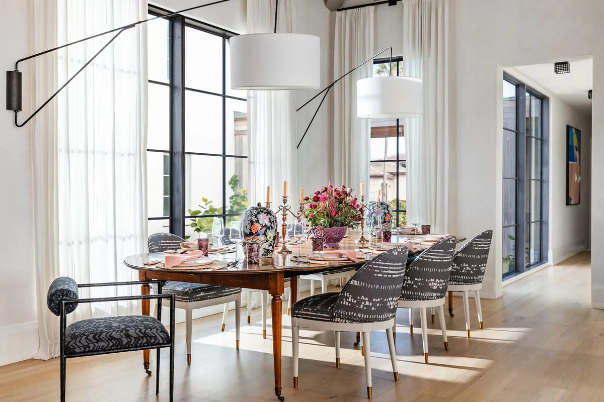 Dining room with a round table set for eight, patterned chairs, flowers, large windows, and a floor length mirror