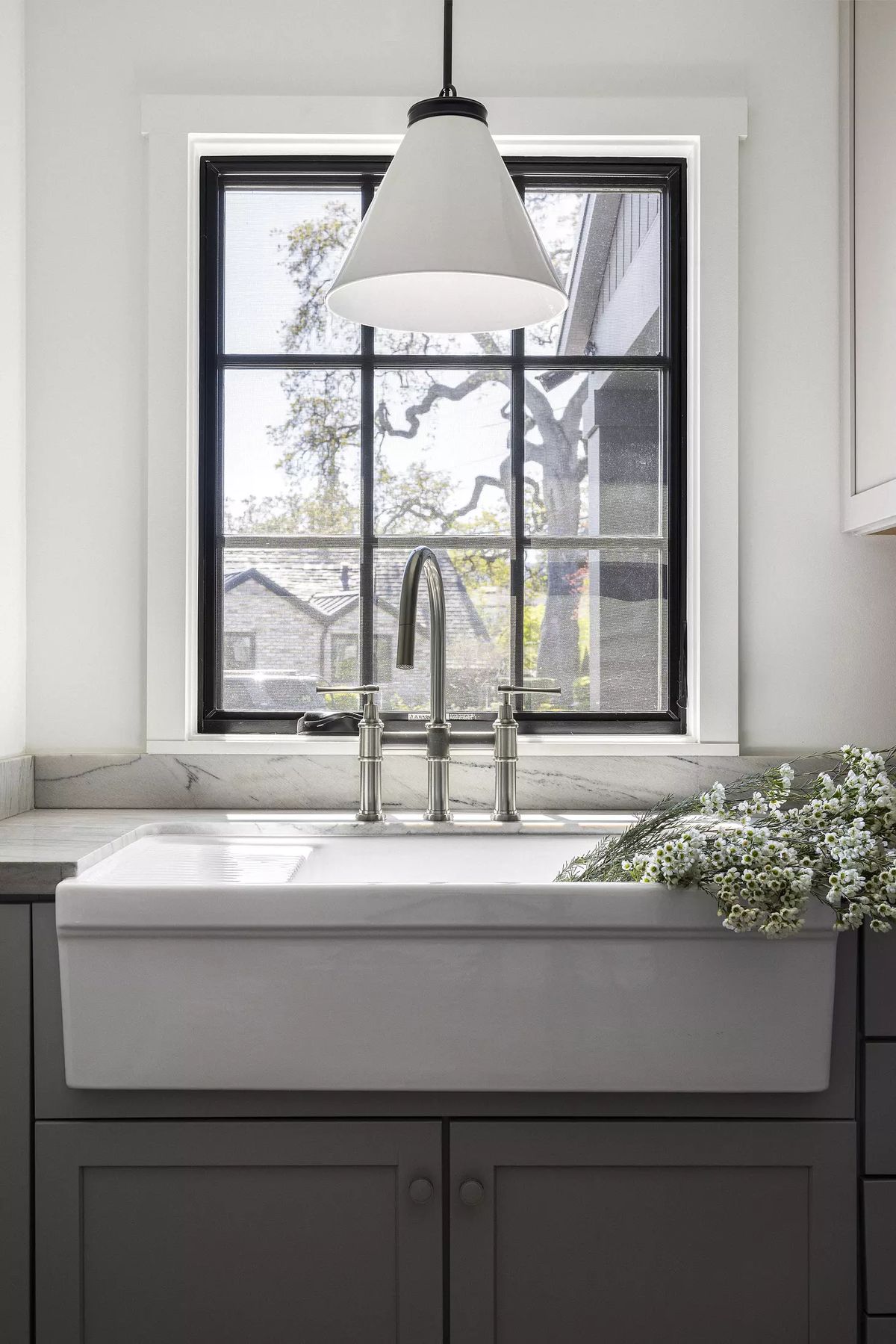 Kitchen with farmhouse sink, gray cabinetry, marble countertop, and large window with white flowers.