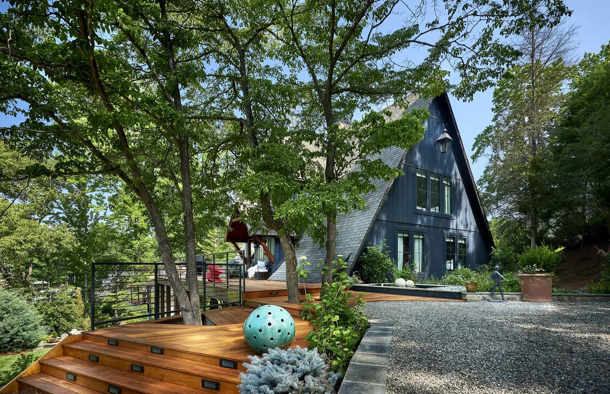 Exterior front view with wooden staircase, dark house facade, trees, shrubs, and decorative elements.