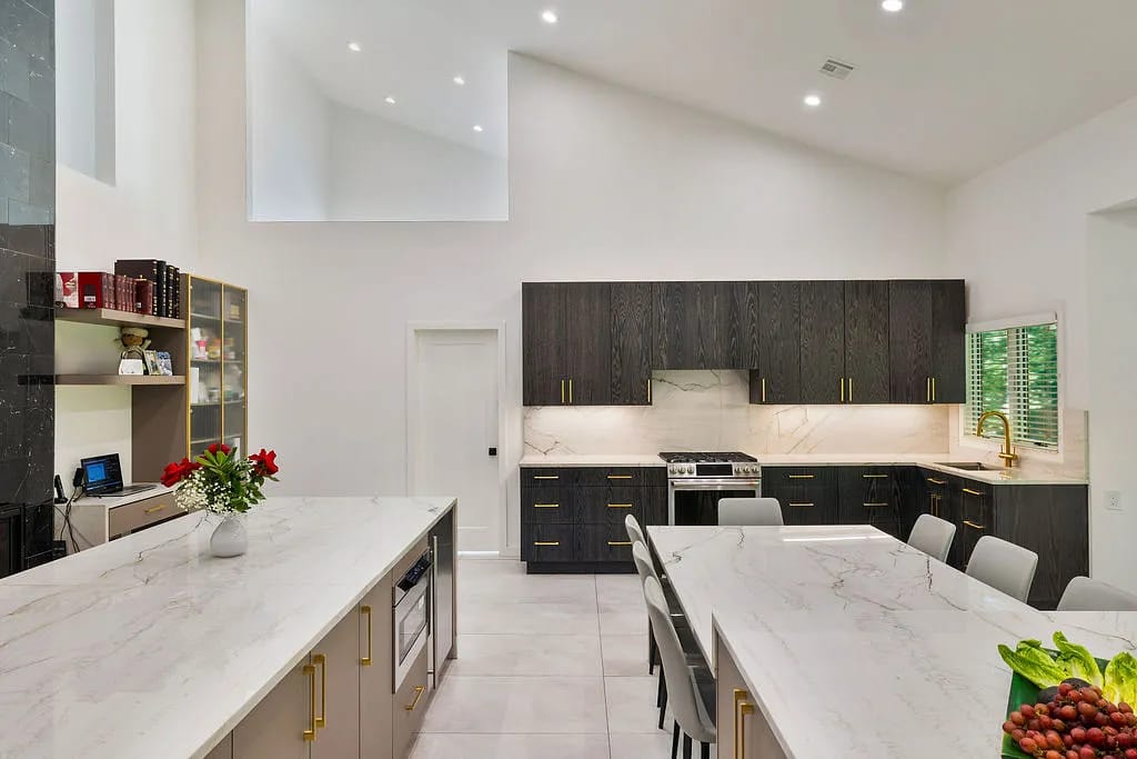 Kitchen with marble island, dark wood cabinets, stainless steel appliances, white chairs, and a high ceiling.