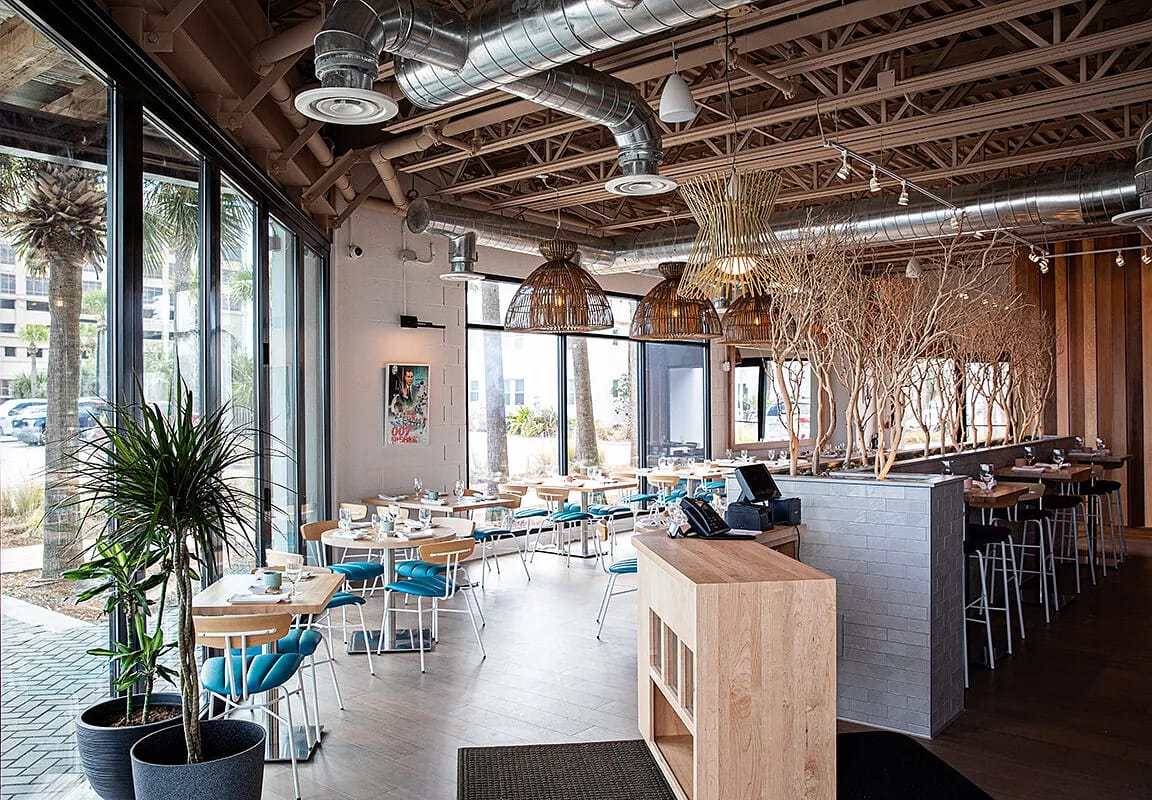 Dining room with round tables, blue chairs, bar area, large windows, exposed beams, and decorative branches.