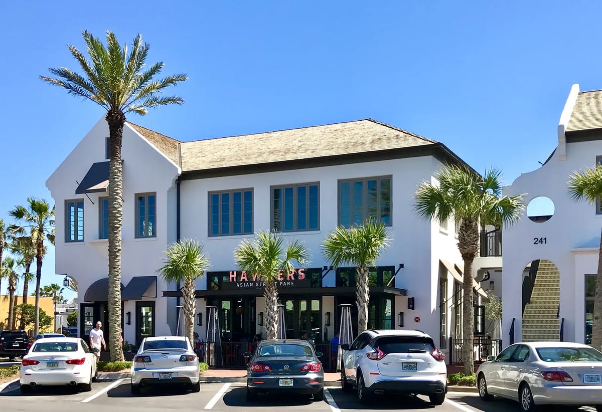 Exterior front of a restaurant with large windows, palm trees, and parked cars.