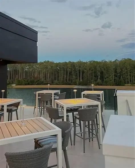 Outdoor deck with white tables, gray chairs, and a view of the lake and trees.