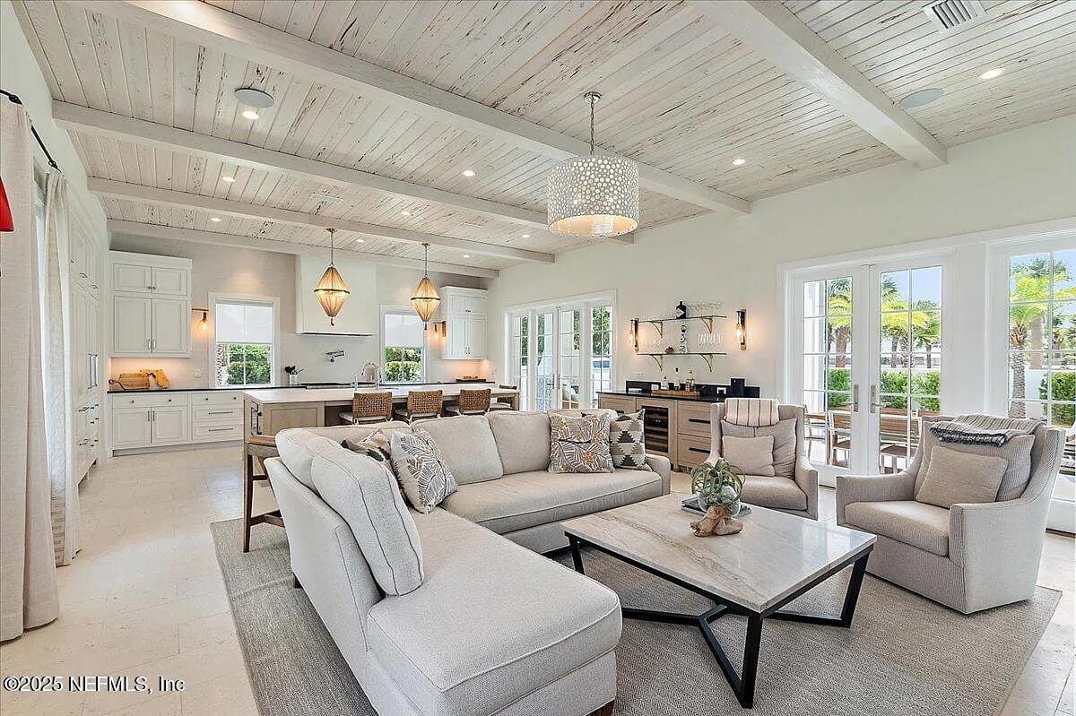 Living room with sectional sofa, coffee table, armchairs, wood beams, large windows, and kitchen view