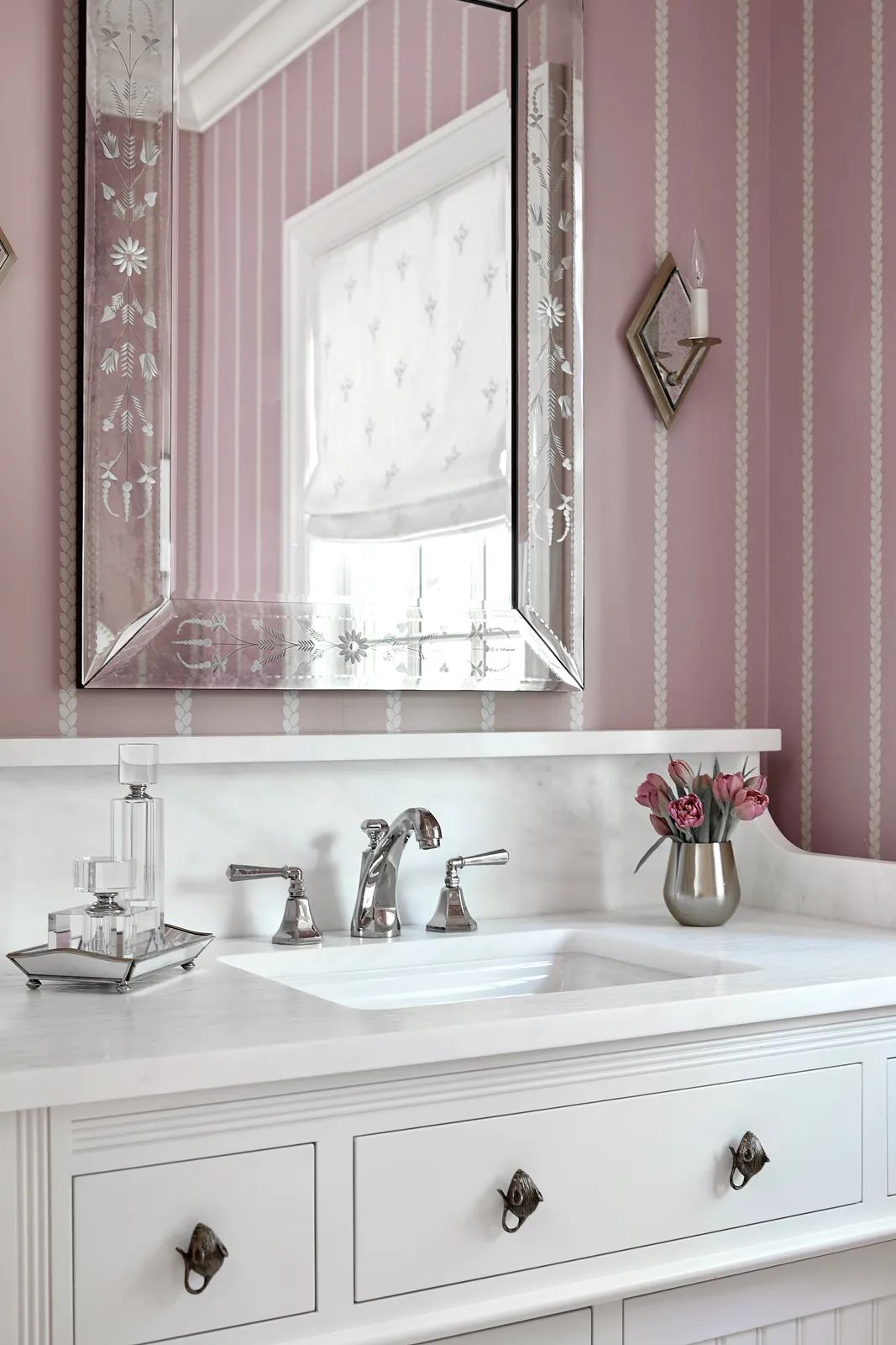 Bathroom with white marble countertop, twin sinks, decorative mirror, pink wallpaper, and floral arrangement.