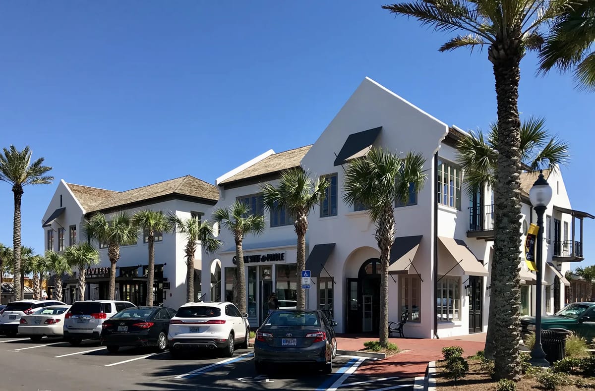 Commercial building with white walls, thatched roof, palm trees, and cars in front.