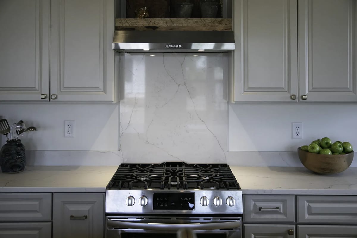 Kitchen with gray cabinetry, white countertops, gas stove, marble backsplash, and wooden bowl of green apples.