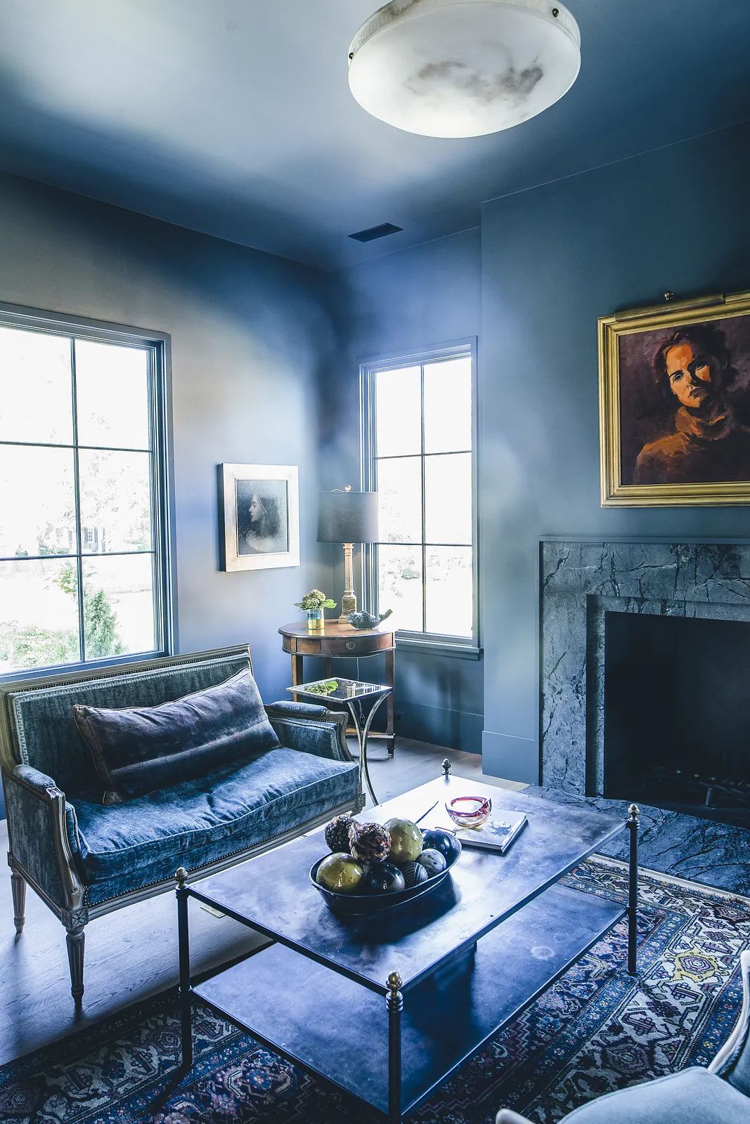 Living room with blue walls, green velvet sofa, glass coffee table, area rug, and marble fireplace with natural light.