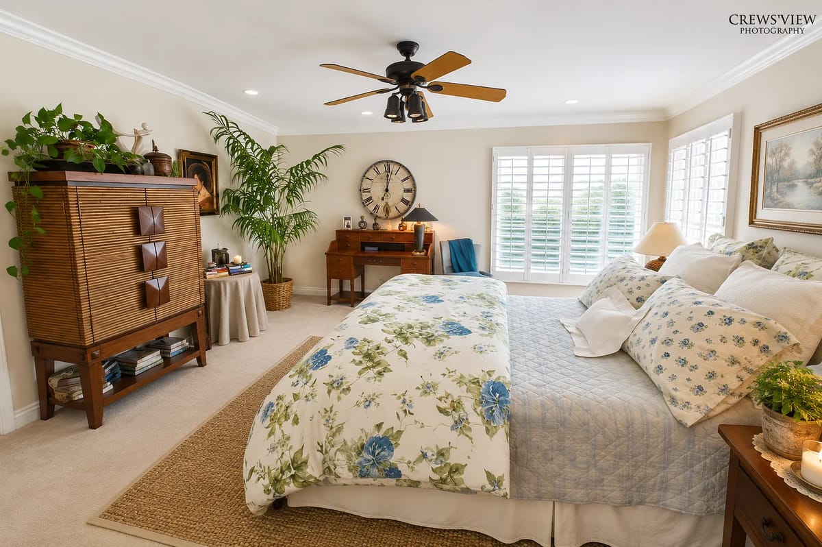 Bedroom with floral quilt, wooden dresser, nightstand, chair, and potted plant with plantation shutters.