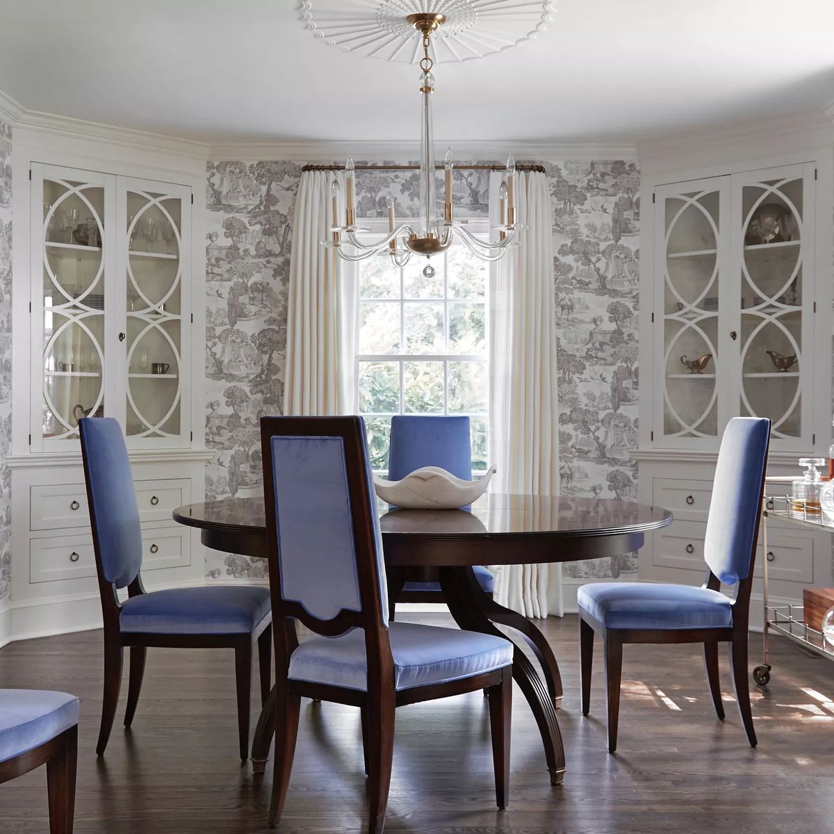 Dining room with round wooden table, blue chairs, glass-front cabinets, and patterned wallpaper.