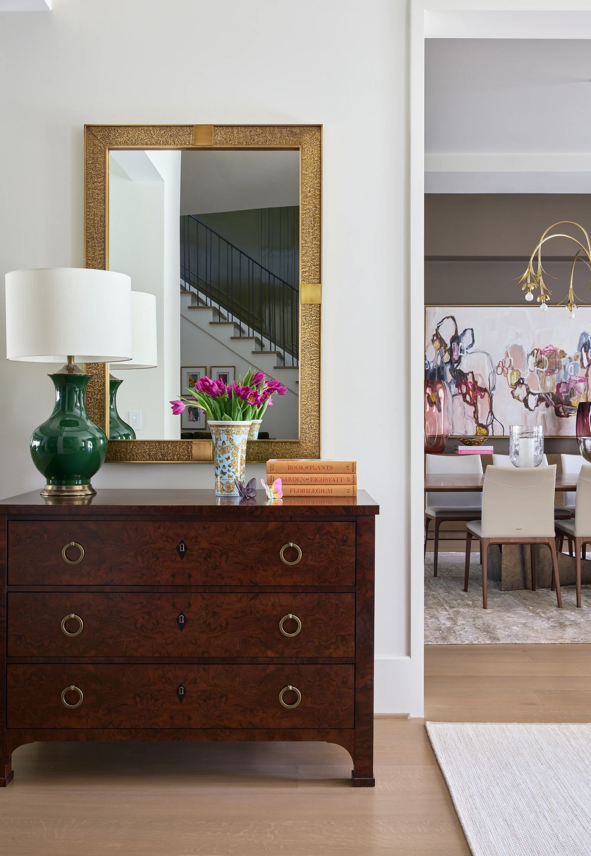 Foyer with dark wood chest, green lamp, flowers, mirror, and visible staircase and dining room.