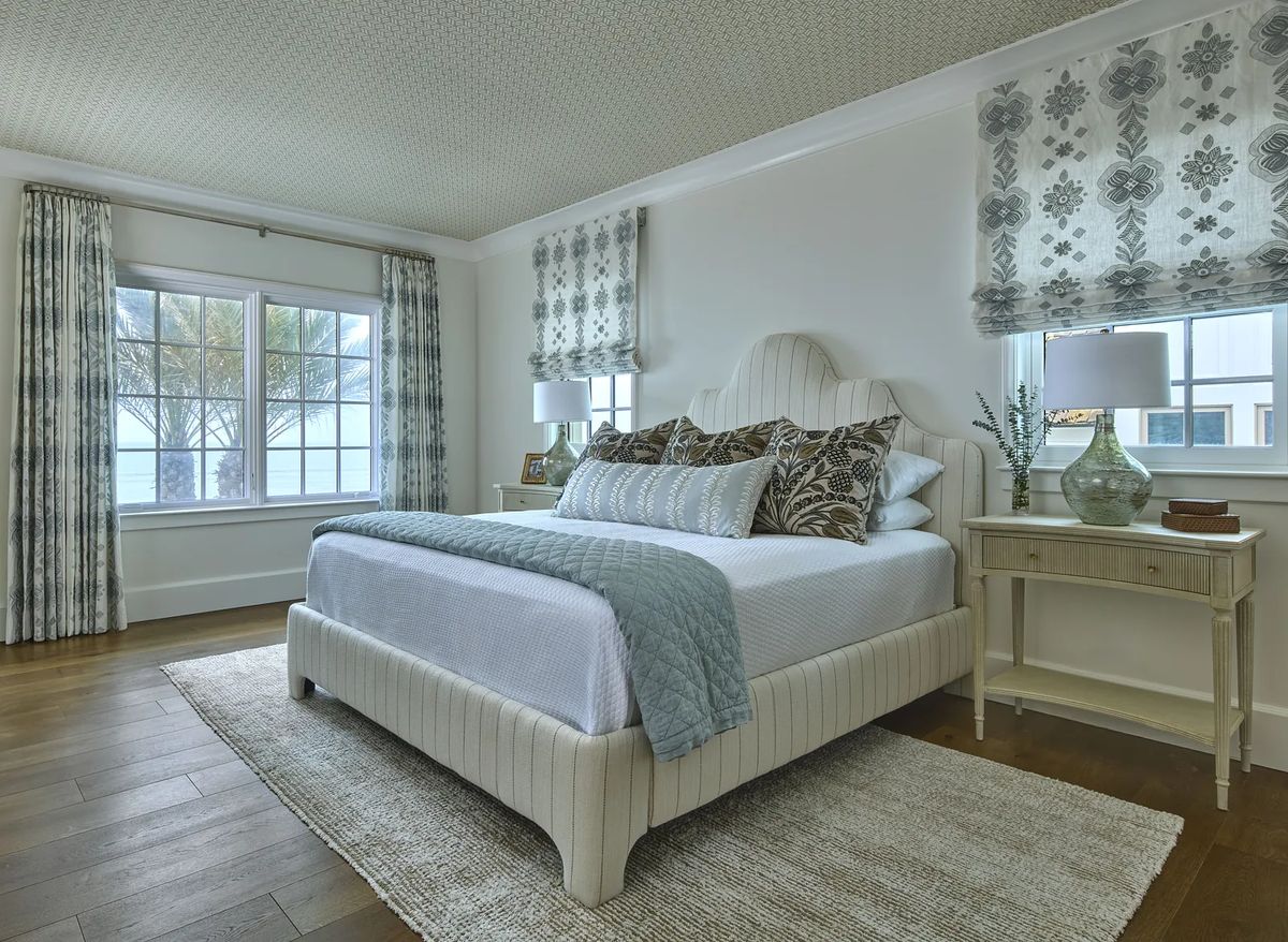 Bedroom with bed, blue quilt, nightstands, lamps, large windows, and patterned rug on hardwood floor