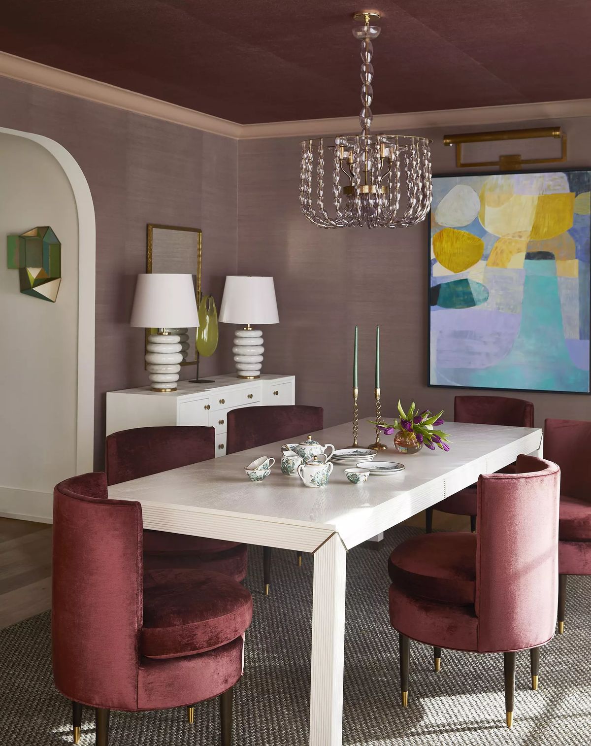 Dining room with white table, burgundy chairs, abstract art, and decorative lamps on sideboard.
