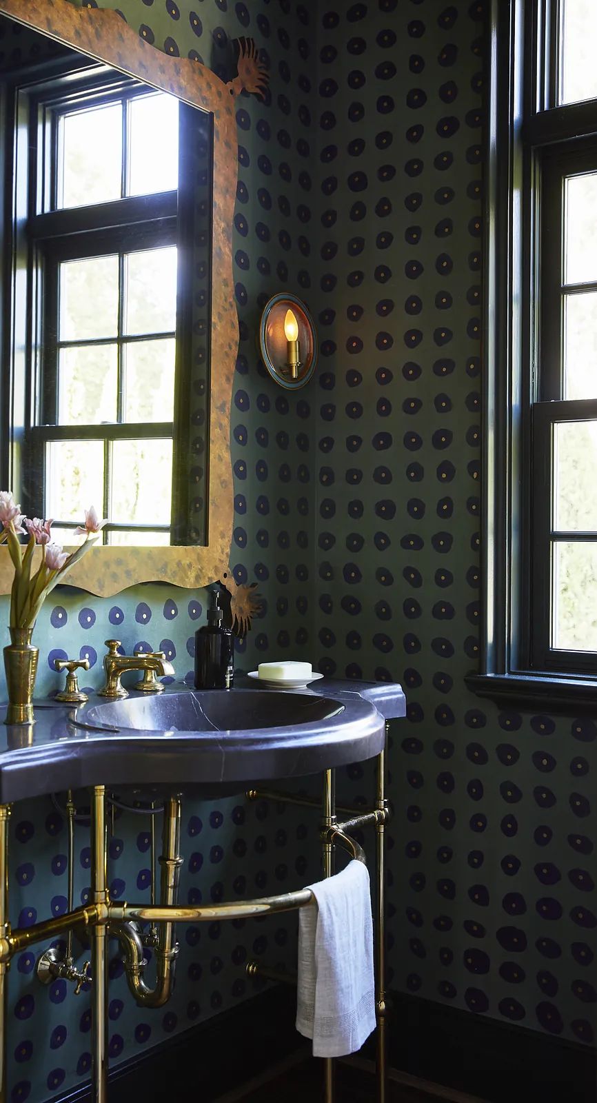 Bathroom with marble sink on brass frame, dark patterned wallpaper, two windows, and flowers in a vase.