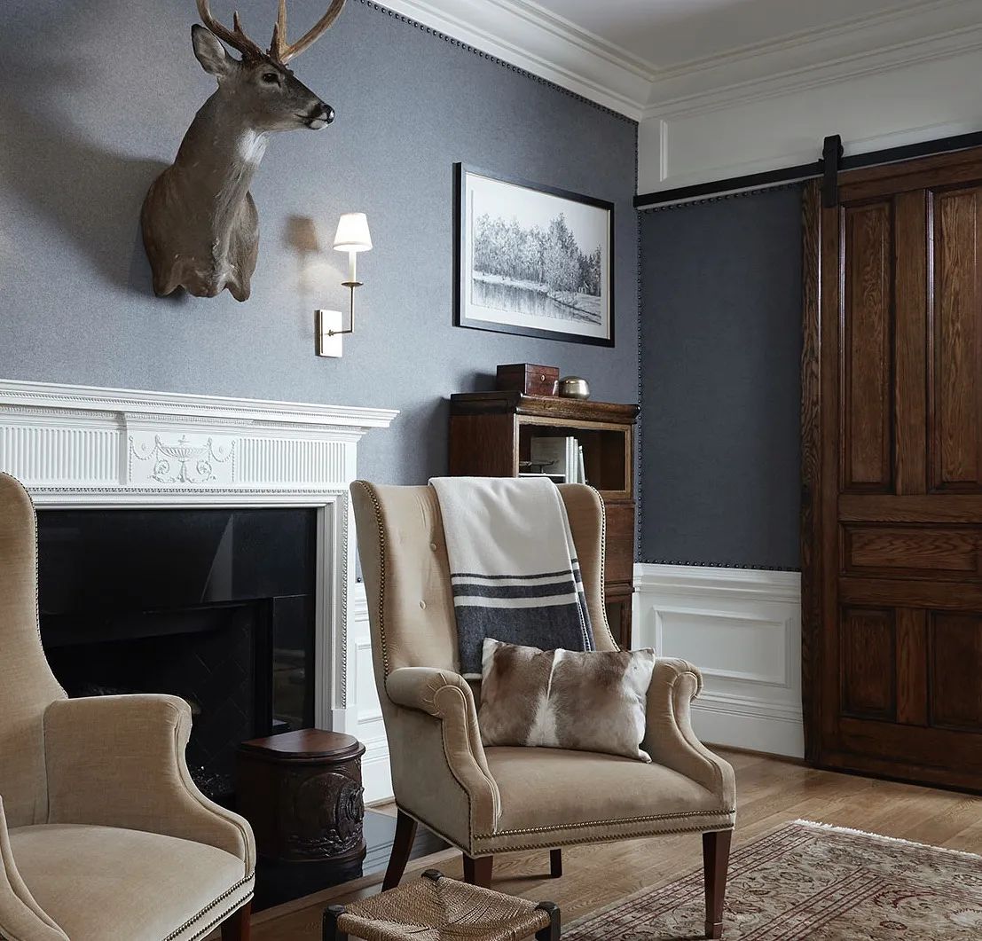 Living room with beige armchairs, round wooden table, fireplace, and mounted deer head.