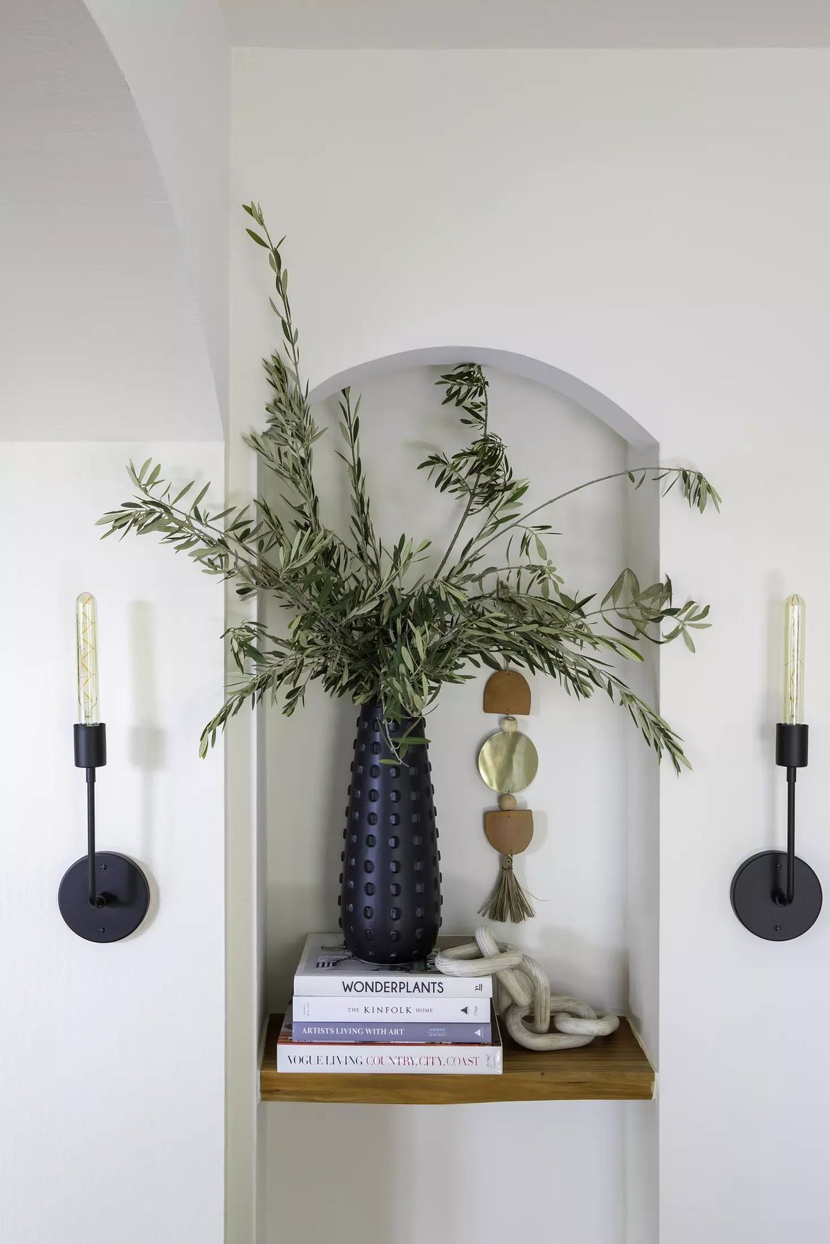 Decorative alcove with black vase, greenery, stacked books, and wall sconces on a light wall.