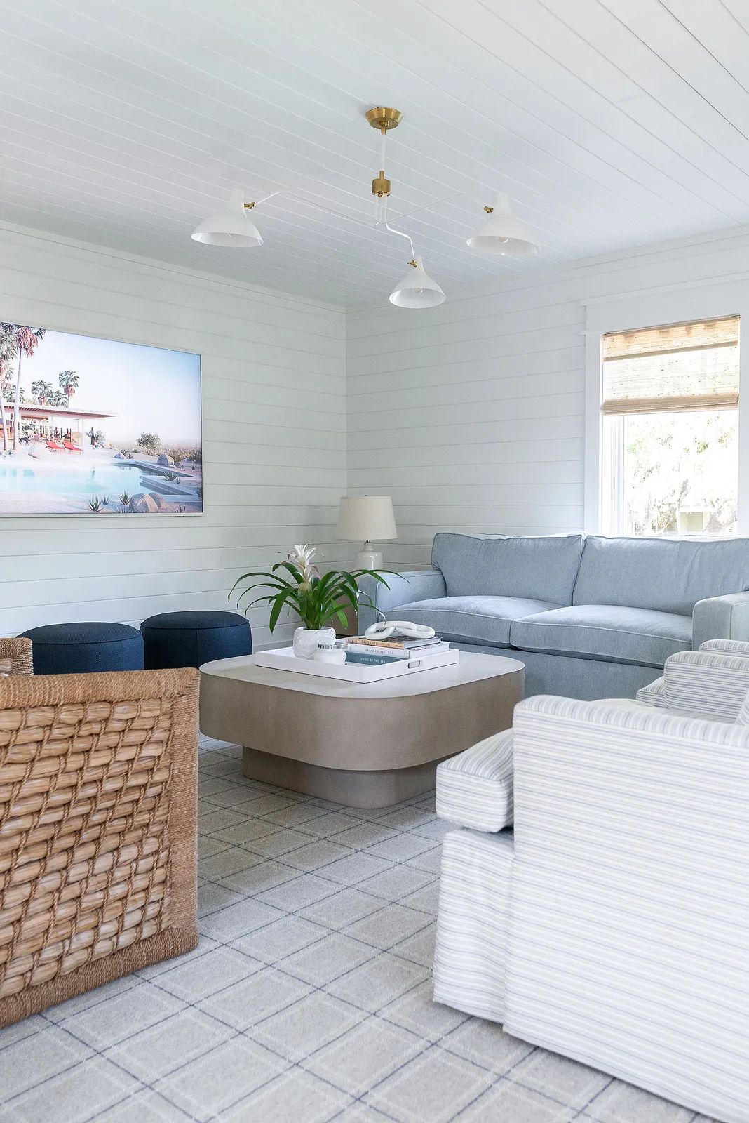 Living room with blue sofa, coffee table, two ottomans, woven chair, and large pool scene artwork.