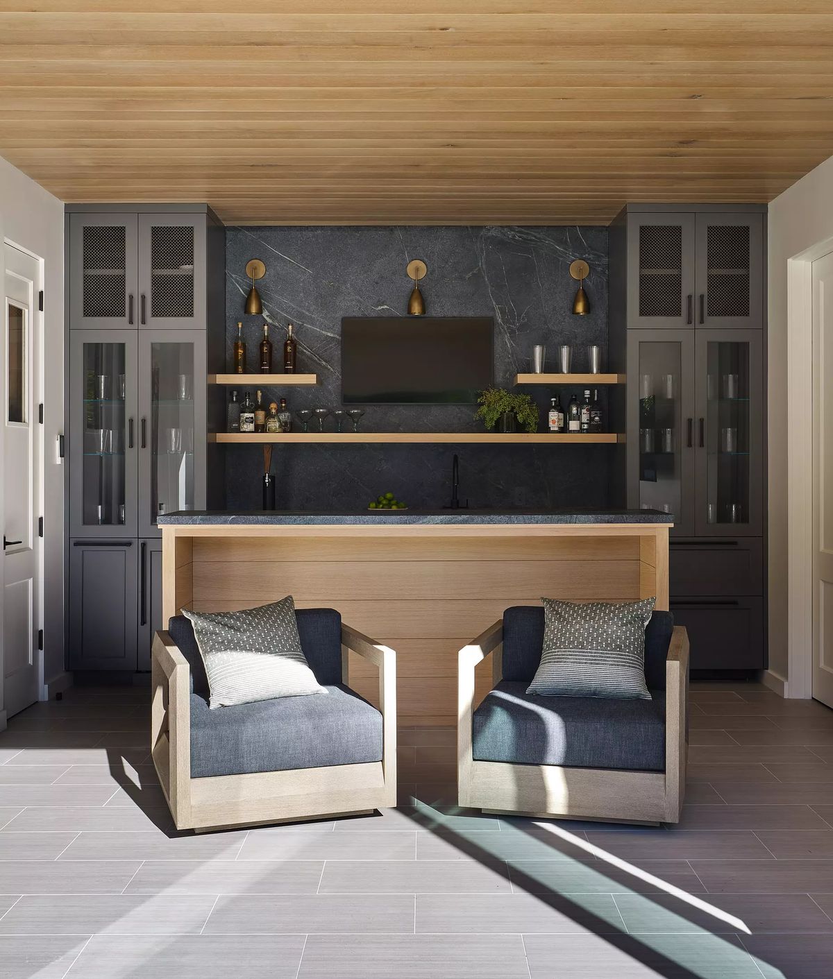 Space with two upholstered chairs, wooden bar, glass cabinets, stone wall, and wall-mounted TV.