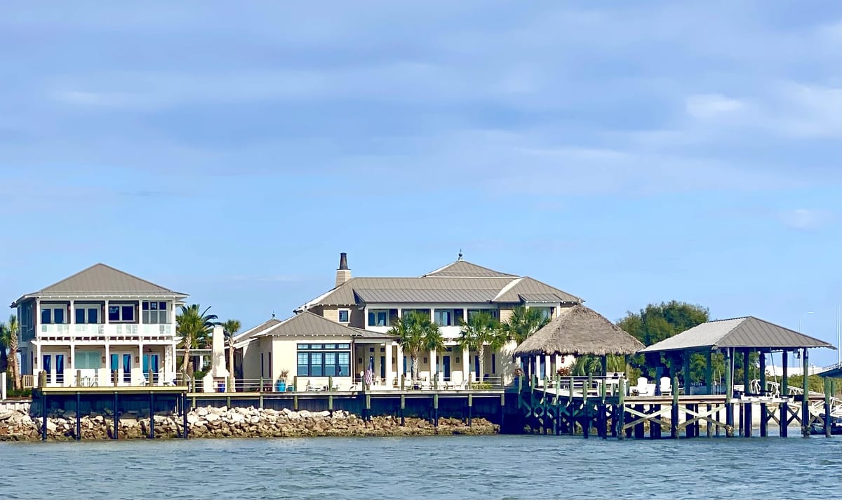 Waterfront exterior with multi-story building, porch, palm trees, and thatched-roof gazebo over a dock.